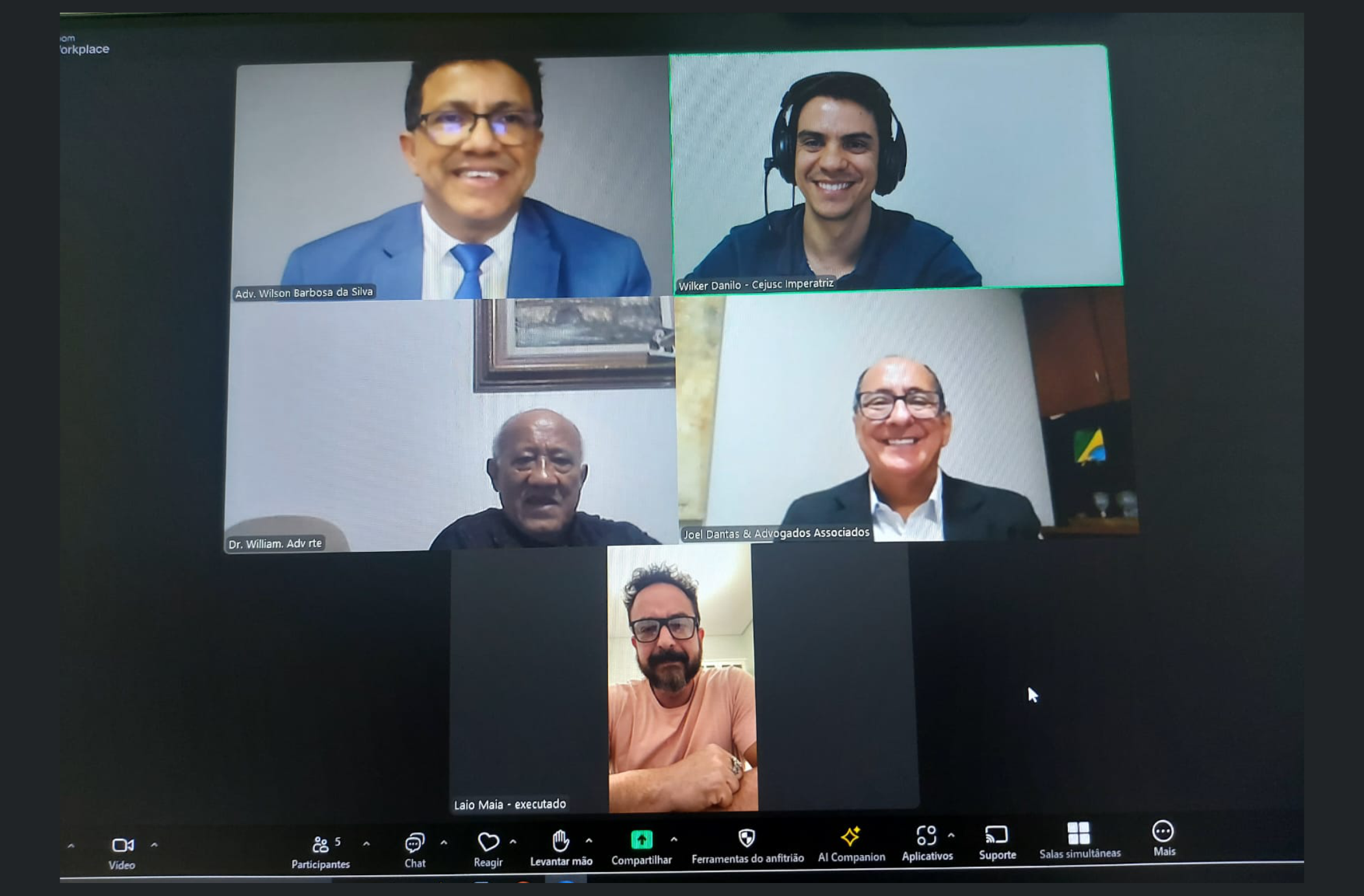 Foto de fundo escuro, mostrando print de uma tela de computador com cinco pessoas sorrindo e posando para a câmera durante realização de uma audiência virtual.