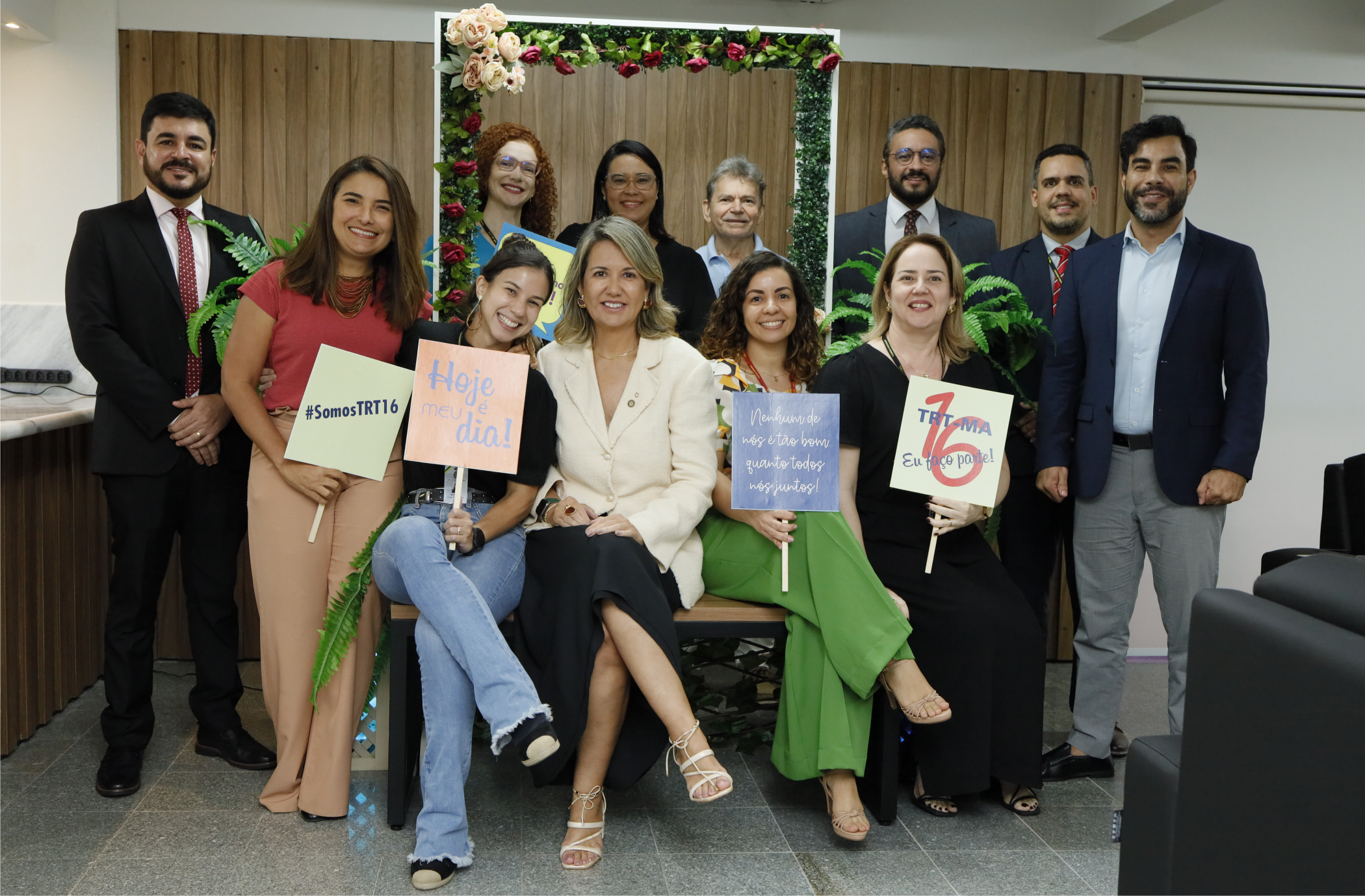 Foto de várias pessoas sorrindo e posando para a câmera, umas sentadas e outras em pé, num cenário decorado com flores.