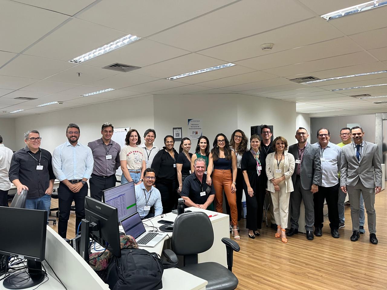 Foto em um escritório com muitas pessoas em pé, sorrindo para a câmera. À frente, mesas com computadores e equipamentos.