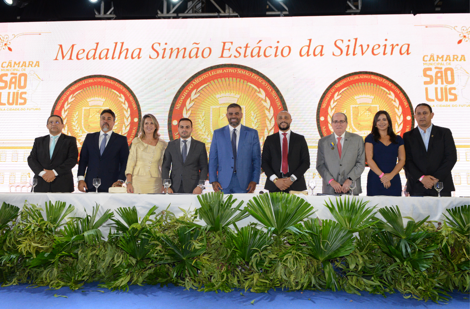  Foto mostra a presidente do TRT-16, desa. Márcia Andrea Farias posicionada na mesa de autoridades com outras personalidades. Na parte de cima, destaque para a frase: Medalha Simão Estácio da Silveira.