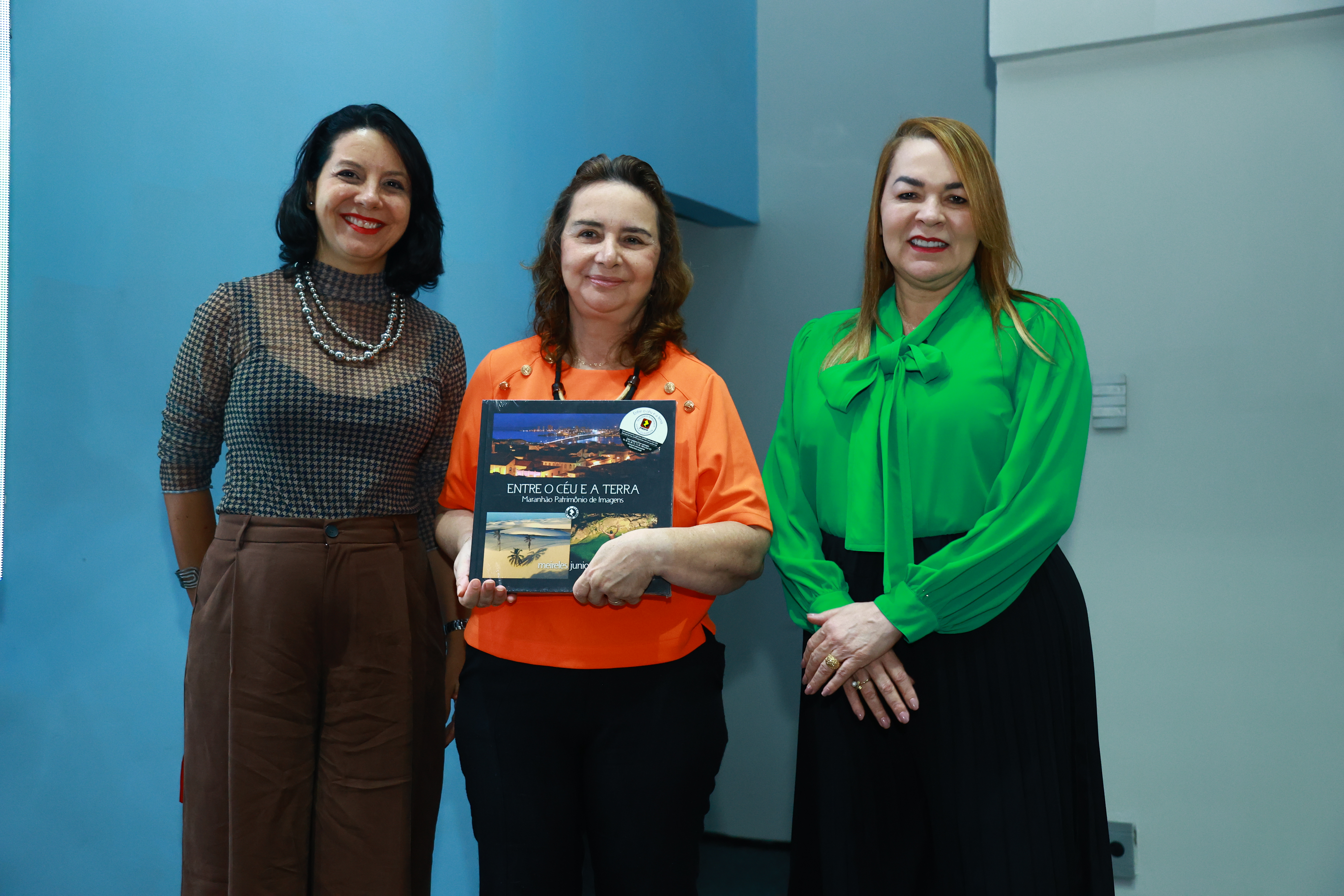 Foto mostra três mulheres, uma ao lado do outra posicionadas para a foto. A que está no centro segura um livro. Ao fundo uma parede azul.