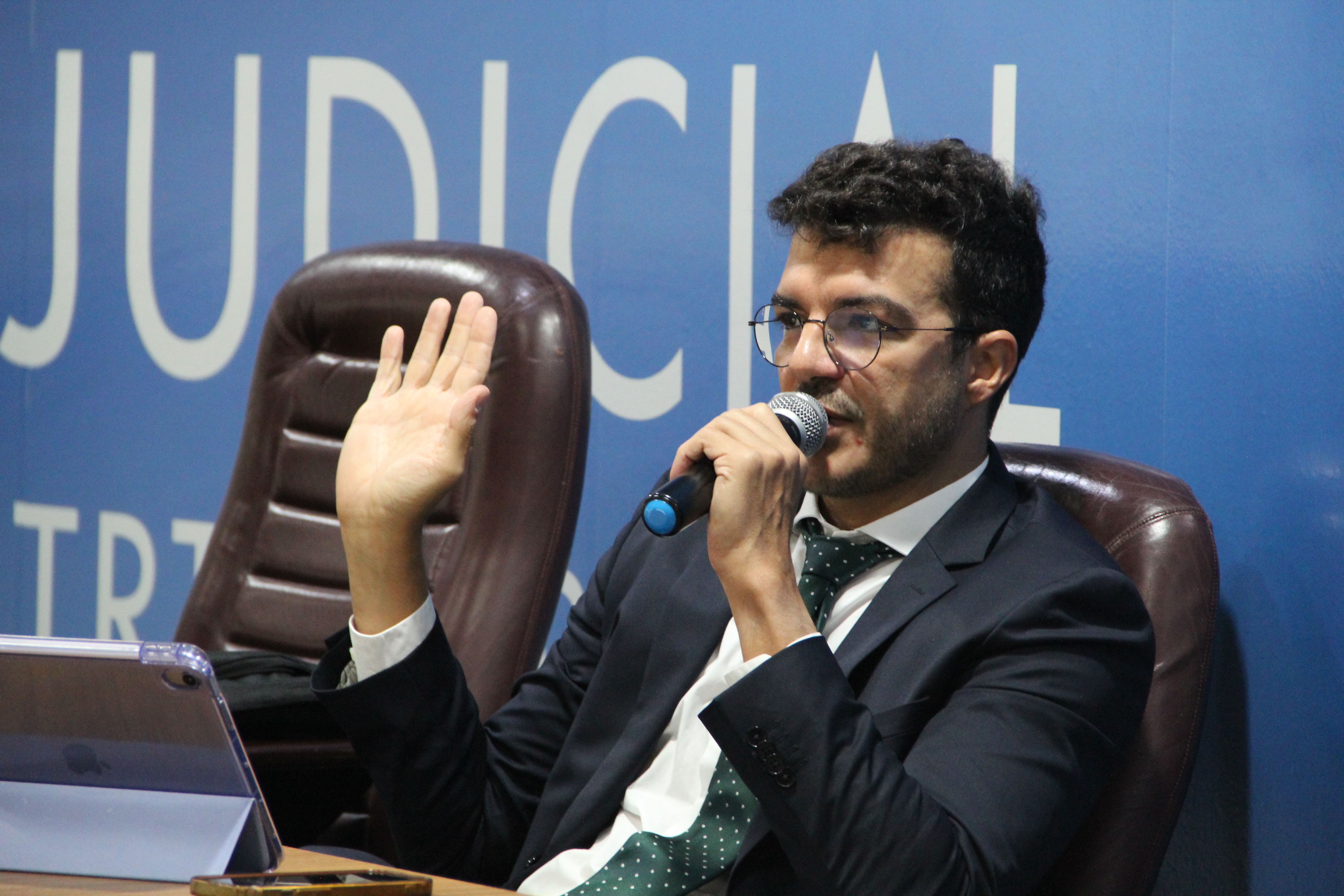 Foto mostra um homem branco usando um terno esculo e camisa azul claro, sentado em uma cadeira. Ele ergue a mão direita e com a mão esquerda segura um microfone. Ao fundo, um painel azul e o nome Escola Judicial.