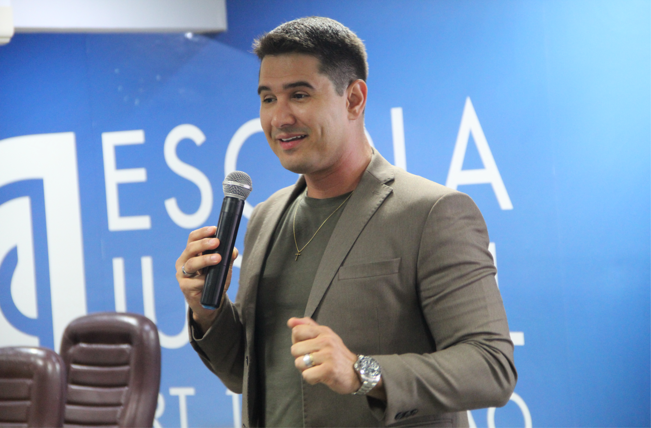 Foto de um homem claro e jovem, usando camisa e blazer caqui. Ele fala ao microfone, que segura com a mão direita. Atrás dele parede azul com inscrição branca. No canto inferior esquerdo, duas cadeiras marrons.