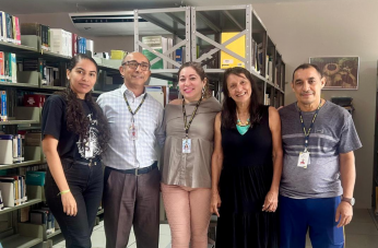 Foto mostra cinco pessoas em pé. Ao fundo prateleiras de alumínio com livros.