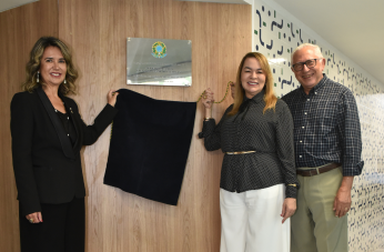Foto mostra duas mulheres e um homem ao lado de uma placa de inauguração posicionada na parede. As duas mulheres 