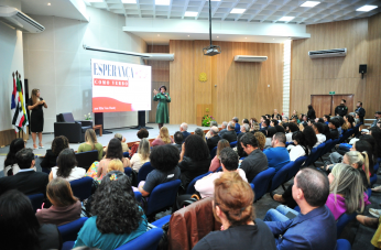 Foto mostra pessoas sentadas em um auditório. No palco, uma mulher se apresenta.