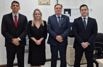 Foto mostra três homens e uma mulher. Todos estão em pé, um ao lado do outro em uma sala. Os homens usam terno escuro e a mulher um vestido longo e escuro.