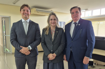 Foto mostra três pessoas, em pé. Sendo dois homens usando terno e uma mulher usando roupa social.