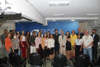 Foto mostra várias pessoas em pé uma ao lado da outra, dentro de um auditório. Ao fundo uma parede azul com no EJUD 16