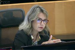 Imagem de uma mulher branca, sentada na cadeira de um plenário. Ela veste blusa estampada e um blazer  na cor preta. 