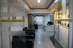 Foto mostra uma a entrada de uma sala. A esquerda tem duas poltronas. Na direita o nome Diretoria Geral. Ao fundo do corredor uma mesa de recepção. 