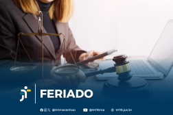 Foto parcial de uma mulher de cabelos claros, usando blazer marrom, segurando um celular na mão esquerda e um martelo de juiz na mão direita, em frente a um notebook aberto. Ao seu lado, vê-se uma balança, representando a Justiça. Em primeiro plano, uma tarja azul com o texto branco FERIADO NA VT ao lado da logomarca do TRT-16. Abaixo do texto, os ícones e endereços do TRT-16 nas redes sociais.