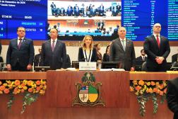 Foto mostra cinco pessoas em pé atrás de uma mesa de honra. Na parte da frente, o brasão do Poder Legislativo