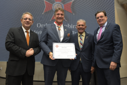 Foto mostra 4 homens em pé, um ao lado do outro. Todos usam termos escursos. Um deles segura nas duas mãos um certificado e usa um colar com uma medalha.