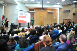 Foto mostra pessoas sentadas em um auditório. No palco, uma mulher se apresenta.