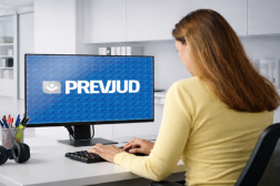 Foto de uma mulher clara de costas, usando blusa de mangas compridas amarela e cabelos longos. Ela está sentada a uma mesa, em frente a um computados, onde se lê na tela PREVJUD.