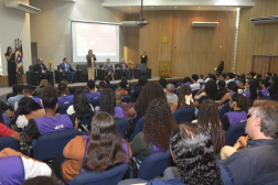 Foto mostra várioas pessoas sentadas em um auditório. Ao fundo, outras seis pessoas estão posicionadas no dispositivo de honra.