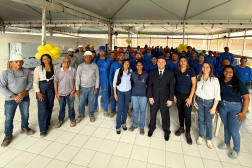 Foto mostra um grupo de pessoas em pé, olhando para a frente. Todos estão em um salão