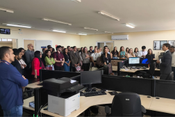 Foto mostra vários pessoas reunidas em pé, dentro de uma grande sala. 