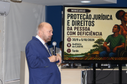 Foto mostra um homem falando ao microfone. Ao lado dele um painel com a informação: Proteção Jurídica e Direito da Pessoa com Deficiência