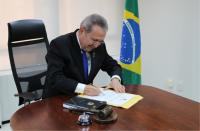 Imagem do desembargador Carvalho Neto sentado em mesa de reunião assinando um documento. Ao lado, a imagem da Bandeira do Brasil em mastro.