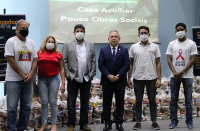 Seis pessoas em pé no palco de auditório tendo ao fundo telão da campanha Corregedoria Solidária e cestas de alimentos.