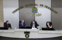 Três homens e uma mulher sentados à mesa do Plenário, à frente de parede listrada de branco e marrom com a inscrição Tribunal Regional do Trabalho 16ª Região em dourado e brasão da República.