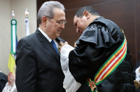 À esquerda, o presidente Carvalho Neto vestido de terno escuro recebe a medalha do desembargador Luis José de Jesus Ribeiro que usa toga e que se encontra à direita e de frente para o homenageado. Ao fundo duas bandeiras em mastros