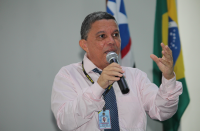 Foto de um homem falando ao microfone. Ao fundo, as bandeiras do Brasil e do Maranhão.