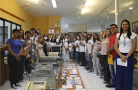 Foto de vários estudantes (moças e rapazes), além de mulheres e homens adultos. Todos estão em pé numa sala ampla, onde estão expostos objetos antigos (máquinas de escrever, computador e telefone) e impressos. 