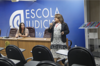 Foto de duas mulheres, uma delas está sentada e a outra está em pé, falando do microfone. Elas estão próximas a uma parede azul que tem a logomarca e o nome da Escola Judicial.