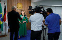Foto de uma mulher clara de frente, usando vestido longo verde e sendo entrevistada por um repórter, que segura um microfone, e dois cinegrafistas operando filmadoras, os três de costas para a câmera.