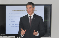 Foto de um homem claro, usando terno e gravata escuros. Ele sorri e aponta para a frente com a mão direita. Atrás dele, uma tela de projeção com textos na cor azul.