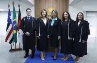 Foto de quatro mulheres e um homem de pé, sorrindo e posando para a câmera. Atrás deles, parede branca com recorte amadeirado onde se vê o brasão da República em dourado. À esquerda, veem-se bandeiras hasteadas.