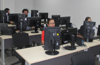 Foto de pessoas sentadas à frente de computadores, tendo ao fundo uma parede branca. Em primeiro plano, uma mulher, usando blusa vermelha e óculos, sorri.