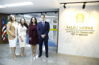 Foto de três mulheres e um homem de pé, sorrindo e posando para a foto. Atrás deles, porta de vidro com bandeiras hasteadas à esquerda. No lado direito da foto vê-se parede bege iluminada e decorada com o brasão da República e texto, ambos dourados, onde se lê SALÃO NOBRE JUSTIÇA DO TRABALHO TRT 16ª REGIÃO.