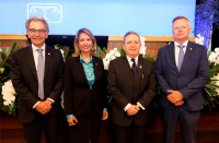 Foto de três homens e uma mulher, todos estão de pé, usam ternos escuros e e sorriem para a câmera. Atrás deles, decoração de folhagem verde com flores brancas, mesa amadeirada e tela de projeção em tons de azul.