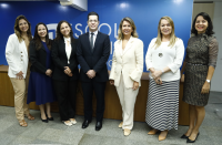 Foto de um homem usando terno escuro ladeado por seis mulheres. Todos estão de pé e posam para a câmera, sorrindo. Atrás deles, parede azul e mesa de madeira.