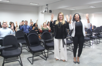 Foto mostra duas mulheres em pé, posicionadas em frente às cadeiras de um auditório. Elas vestem roupas sociais nas cores preto e branco. Ao fundo pessoas diversas sentadas. Todas estão com as mãos erguidas para cima, fazendo um sinal em Libras. 