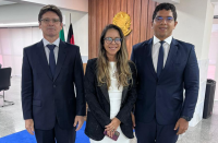 Foto mostra no primeiro plano e no centro uma mulher usando roupas sociais nas cores branco e preto, ela tem cabelos longos, usa óculos. Ela está ladeada por dois homens que usam ternos escuros, com camisas brancas e gravatas no tom azul.