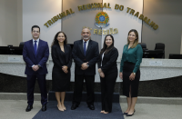 Foto de dois homens e três mulheres de pé, posando para a câmera e sorrindo. Atrás deles, parede em tons de branco e bege com texto dourado e brasão da República, além de bancada de granito preto e mármore branco.