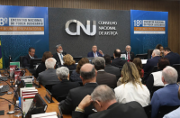 Foto panorâmica de auditório com quatro homens sentados à mesa de honra. Na frente deles, pessoas na plateia. Atrás deles, vê-se parede em tons de cinza e azul com a logomarca do CNJ centralizada. Nas laterais da parede, ilustração com o nome do Encontro Nacional do Poder Judiciário.