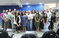 Foto panorâmica de auditório com 22 pessoas, entre homens e mulheres posicionadas em pé. Atrás delas, vê-se uma parede em tom azul.