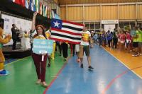 Uma pessoa segurando uma placa que identifica a delegação do TRT-16. Ao fundo, atletas seguram a bandeira do Maranhão
