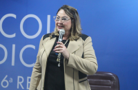 Foto de mulher loira, usando óculos e sorrindo enquanto fala ao microfone, que segura com a mão esquerda. Ela usa blusa preta e blaser bege com detalhes pretos. Atrás dela, vê-se parede azul com inscrição branca e espaldar de uma cadeira marrom.
