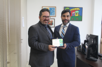 Imagem mostra dois homens no centro, ambos vestidos com terno. Eles seguram uma placa de homenagem. Ao fundo parede branca decorada com quadros coloridos.