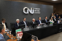 A imagem mostra ao fundo a logomarca do CNJ - Conselho Nacional de Justiça. No primeiro plano, estão posicionados atrás da mesa de honra, autoridades. Ao centro da imagem, aparece sentado o Presidente da República, Luís Inácio Lula da Silva ao lado do presidente do CNJ e do STF, ministro Luís Roberto Barroso.