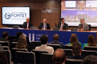 A foto mostra quatro pessoas sentadas à mesa de honra, diante de pessoas em um auditório
