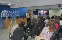 Foro panorâmica de auditório, mostrando a palestrante de frente e a plateia de costas. Ao fundo parede azul, onde se lê ESCOLA JUDICIAL em letras brancas, e tela de projeção com imagem colorida.
