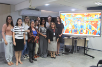 Foto de dez mulheres de pé e sorrindo para a câmera. À sua esquerda, tela de projeção mostrando quadro colorido em tons de laranja e azul.